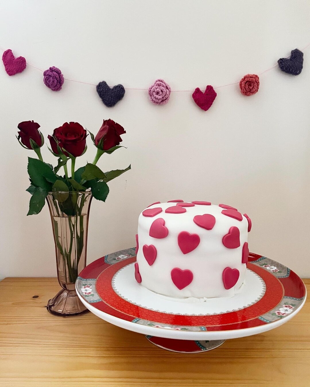 A picture of a decorated cake covered in hearts on a cake stand. On the left, there’s a pink vase with red roses. In the background, against the white wall, hangs a guirlande of knitted pink and purple hearts and roses.