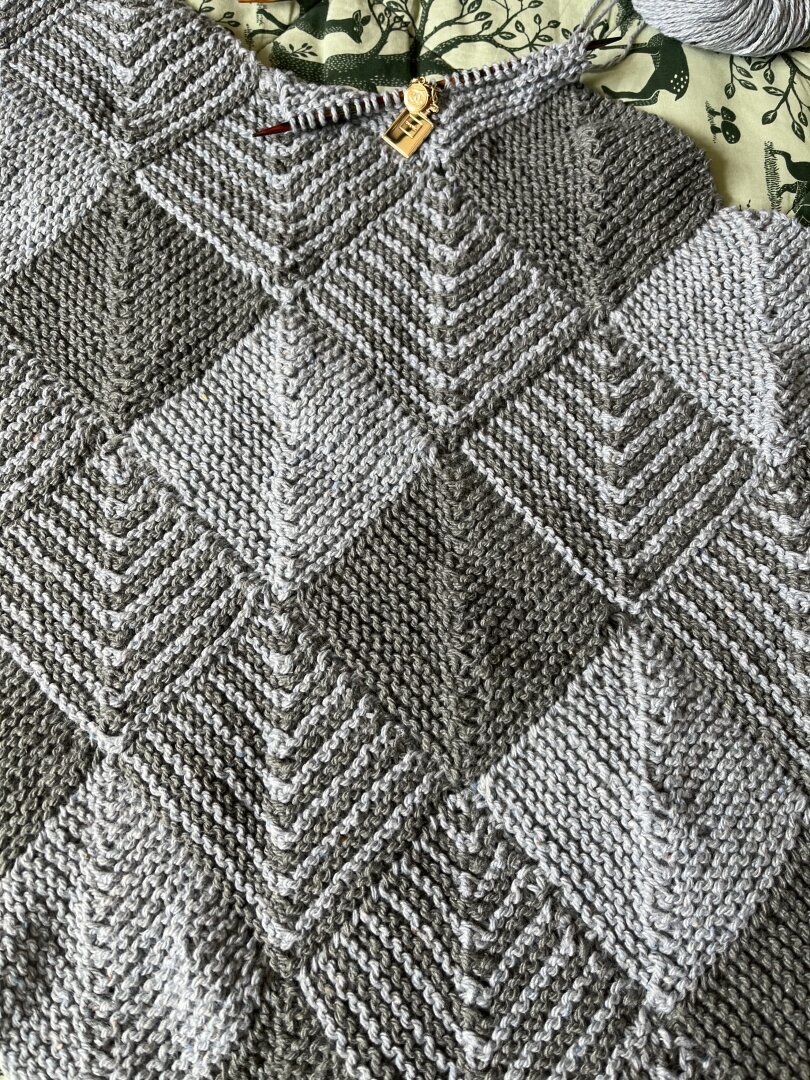 A picture of a knitted blanket in progress. The blanket consist of little squares in light blue and light grey. Some of the squares have both colours, some are a solid colour. At the top of the picture, a square in progress is visible complete with knitting needle and stitch marker