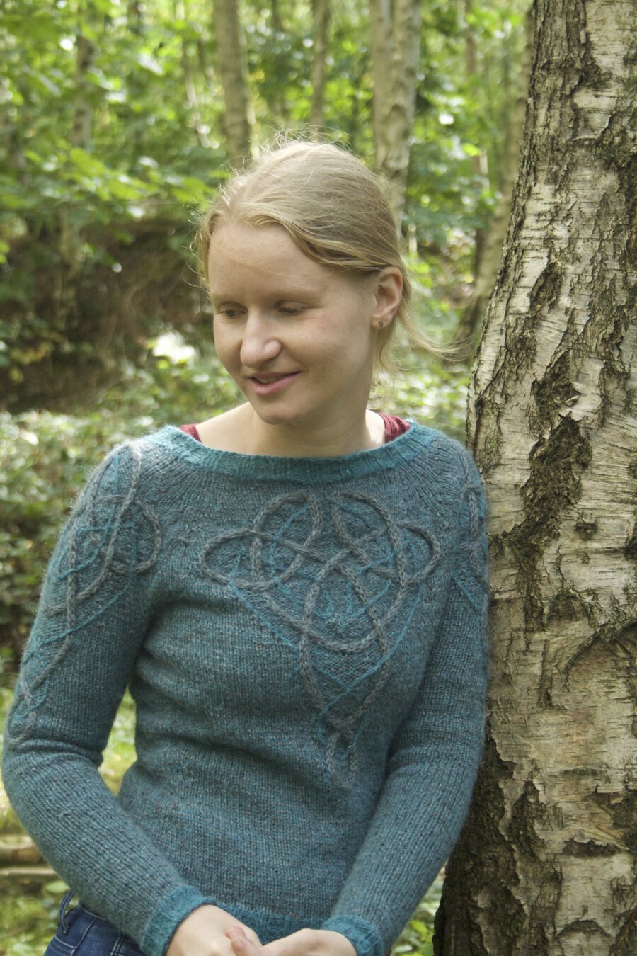 A blonde woman (me) leaning against a tree in a forest. The woman is wearing a knitted blue jumper with a Celtic inspired cable pattern on the front