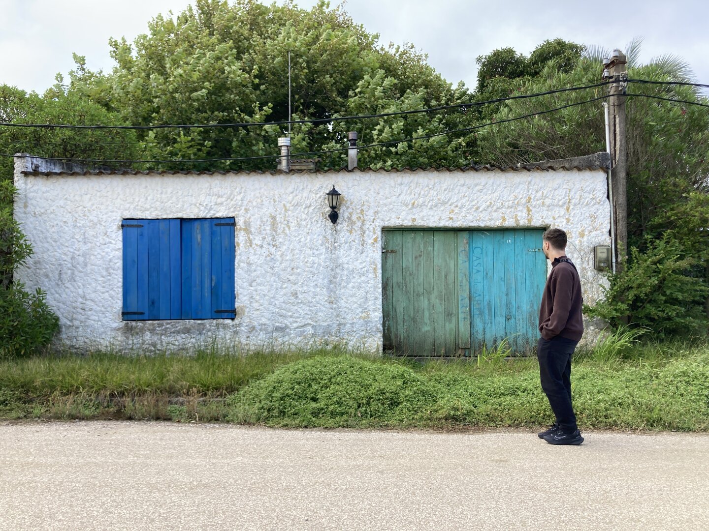 Una fachada blanca con une ventana azul y un portón celeste, los dos de madera 
Delante hay césped y calle de pedregullo . Detrás del rectángulo de la fachada se ven árboles verdes 
Hay un joven caminando