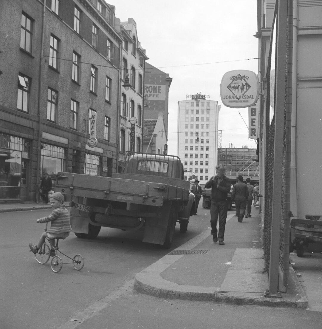 A Bergen black and white cityscape. A flatbed truck is parked on the side of the road. Just behind it a young boy on a trike is ascertaining the traffic to see if he could pass it safely.