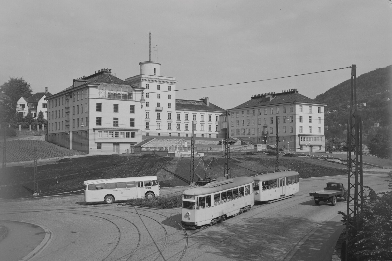 A black and white photo of a large white building with a central round tower and two wings. Between the wings, a greenhouse. At the bottom of the hill, a bus and a tram goes about its day.