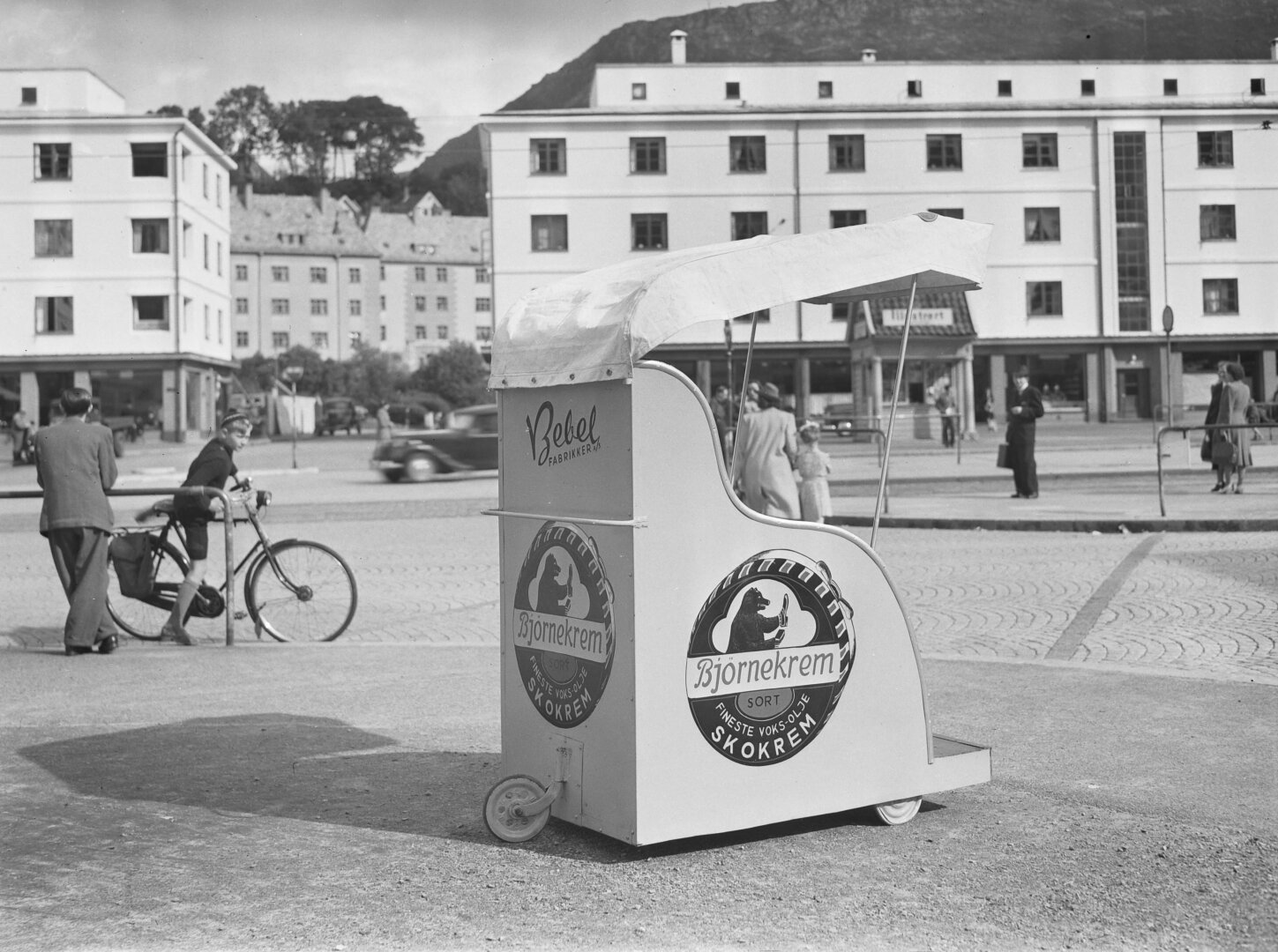 A black and white image of a small roofed shoe polish stand in a city square.