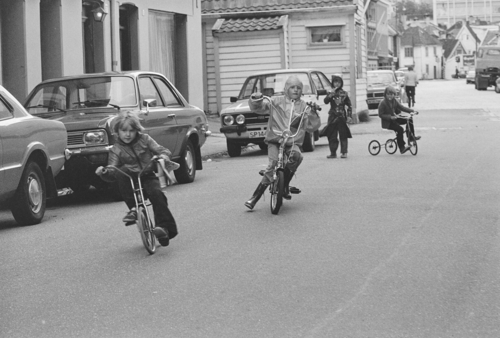 A black and white picture of a 70s street of old wooden buildings with parked cars on the curb. A group of kids are joyriding bicycles in the middle of the street.