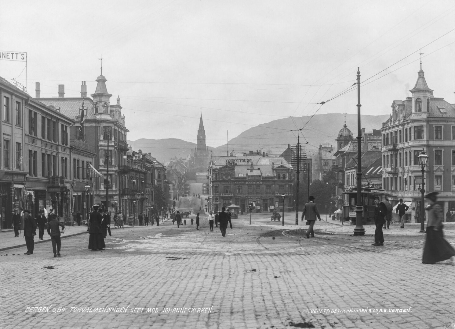 Black and white photo of a large town square in a city, rows of three-story buildings with businesses on the left and right and center, and hilly street going up towards a church tower. People walking on the street, a tram turning towards the right-hand side. Signs read 