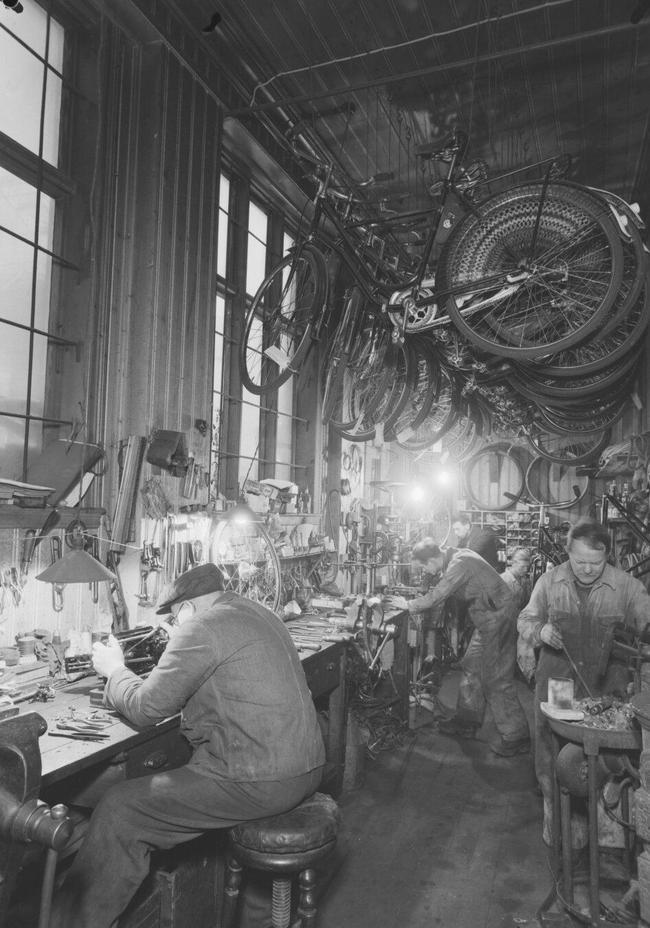 A black and white photograph of a bike workshop. Men working on bike pieces, fine work under desk lamps. In the roof, a number of finished bikes are hanging.