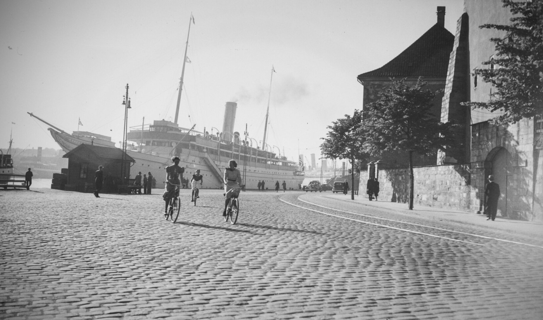 A black and white picture of three women in dresses cycling on a sunny cobblestone street. In the background, a white cruise ship and people walking on the harbour.