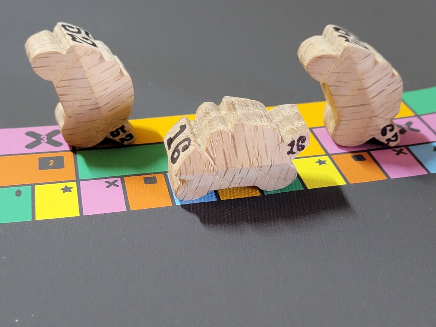 Three hand-carved wooden motorbike game pieces on a colourful track. Two of the pieces are standing on their tail end to look like they are in a wheelie