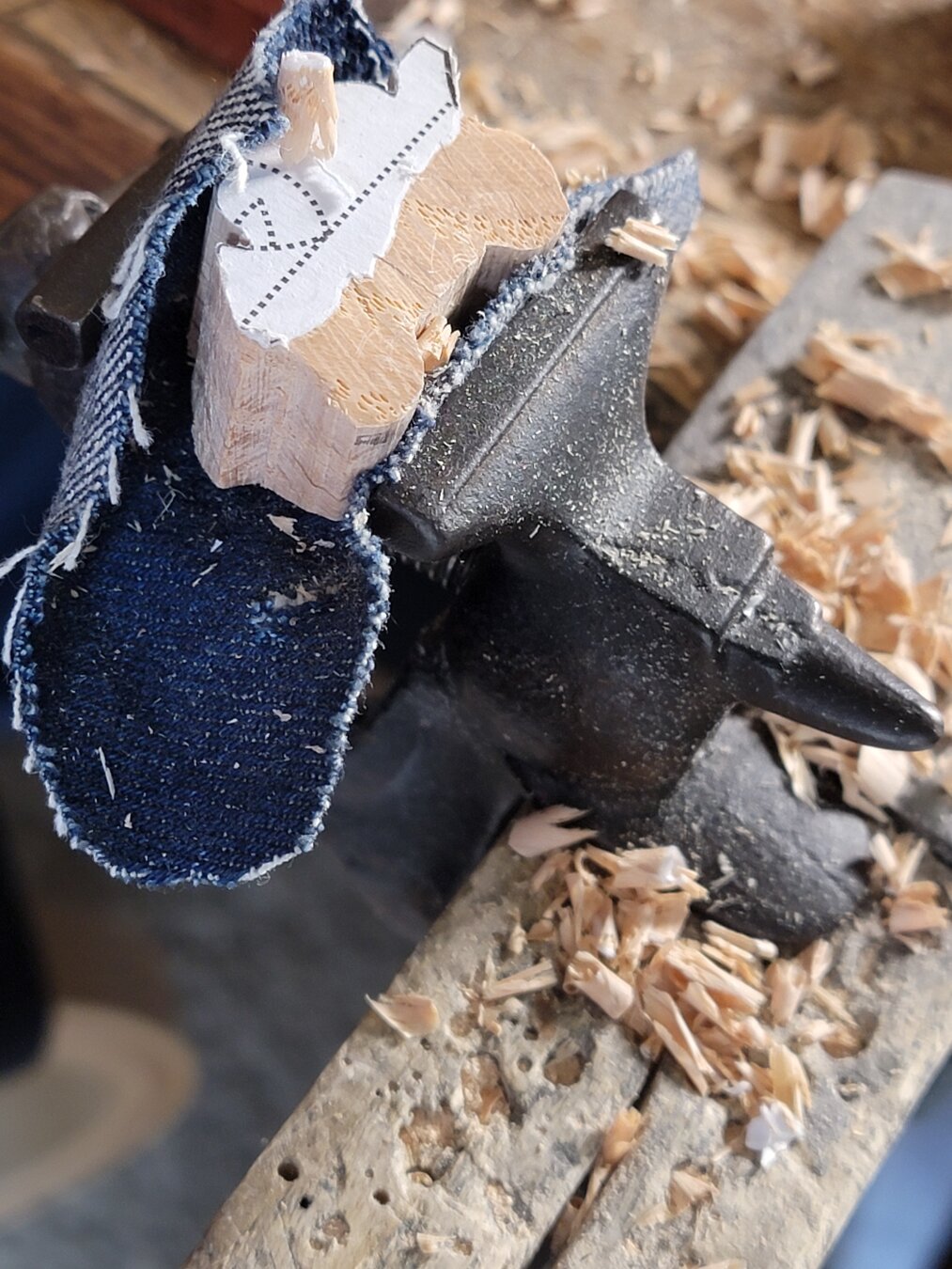 A piece of wood being carved to resemble a motorcycle with rider. The wood is clamped in an antique vice and surrounded by wood shavings