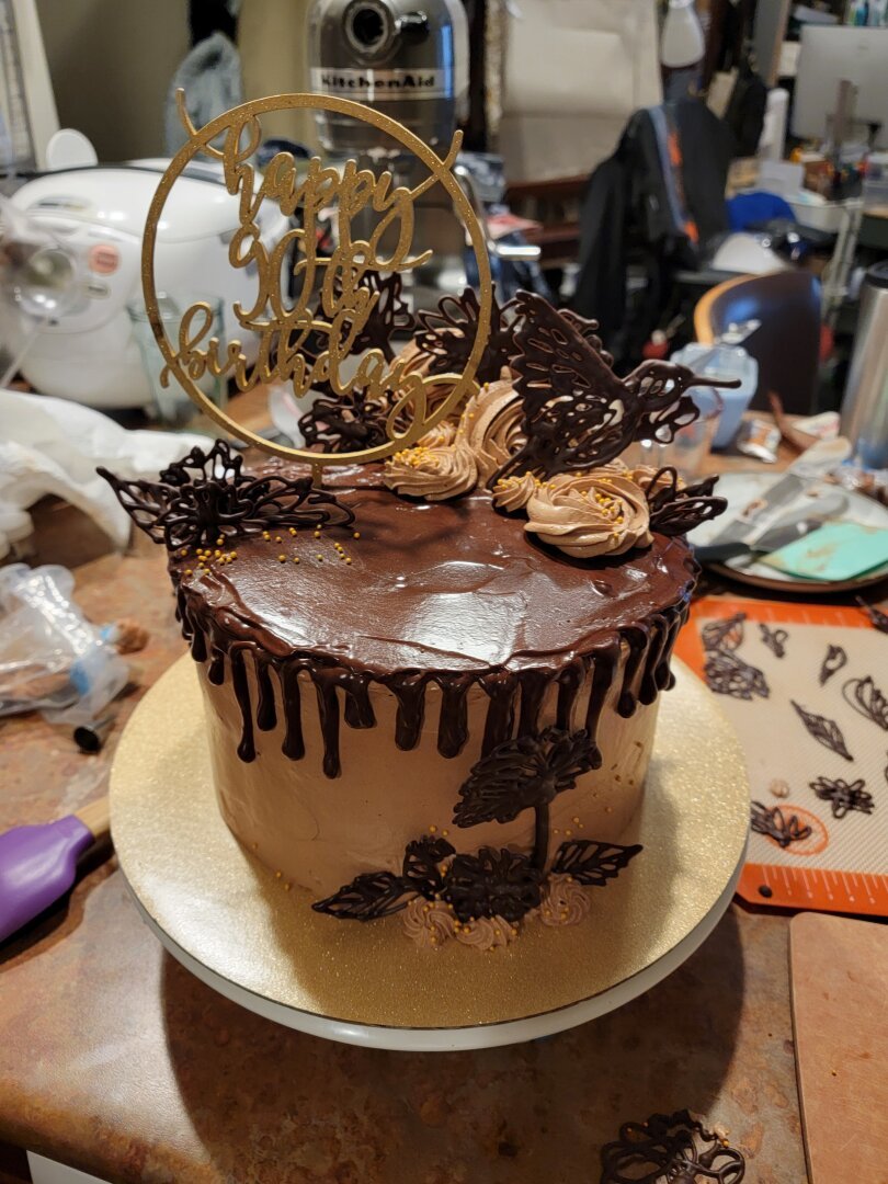 A three-layer, one tier chocolate cake with dark chocolate humming bird and flower decorations, chocolate swiss meringue buttercream, and a dark chocolate ganache drizzle. The cake sits on a glittery, gold cakeboard in a messy kitchen.