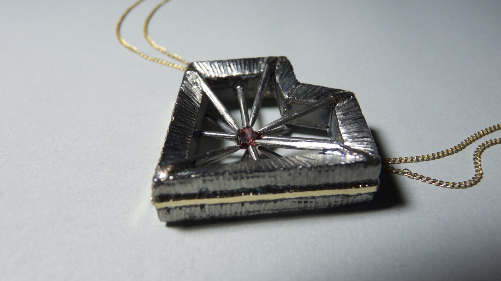 A heart-shaped titanium pendant sits on a white surface. A gold chain trails away from it. The pendant features long pins that point toward the hollow centre of the heart shape, where the pins hold a heart-shaped rhodolite stone.