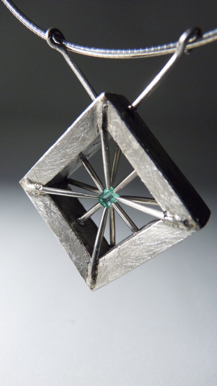 A square pendant hanging at a forty-five degree angle to appear diamond-shaped. The titanium pendant has long pins that all point toward the middle and supports a small emerald.