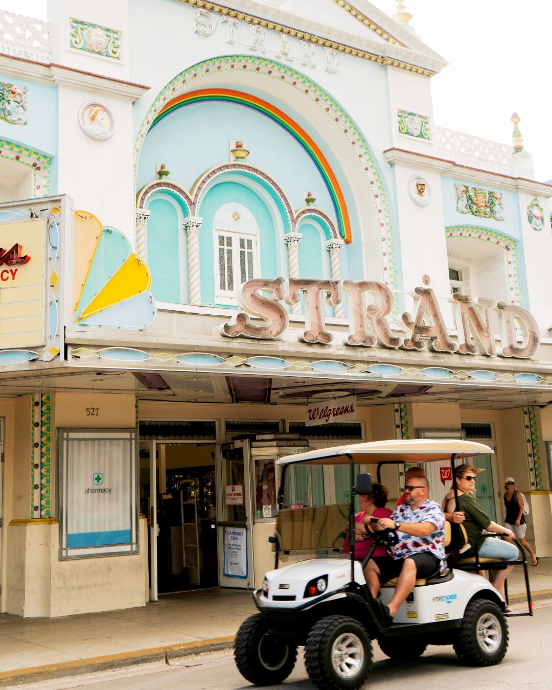 Golf cart riding by with the Strand movie theatre behind it in Key West. A painted rainbow and historical architecture are presented in the background.