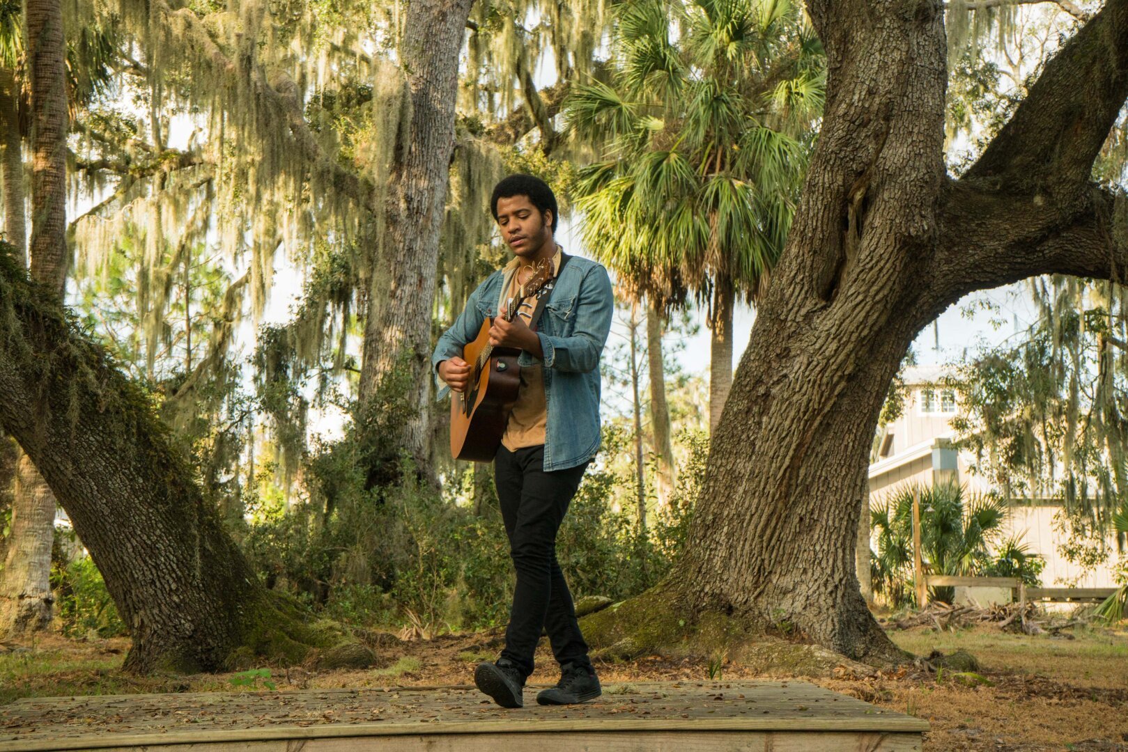 Folk musician sings and plays an acoustic guitar on a wooden stage in the middle of a wooded park area.