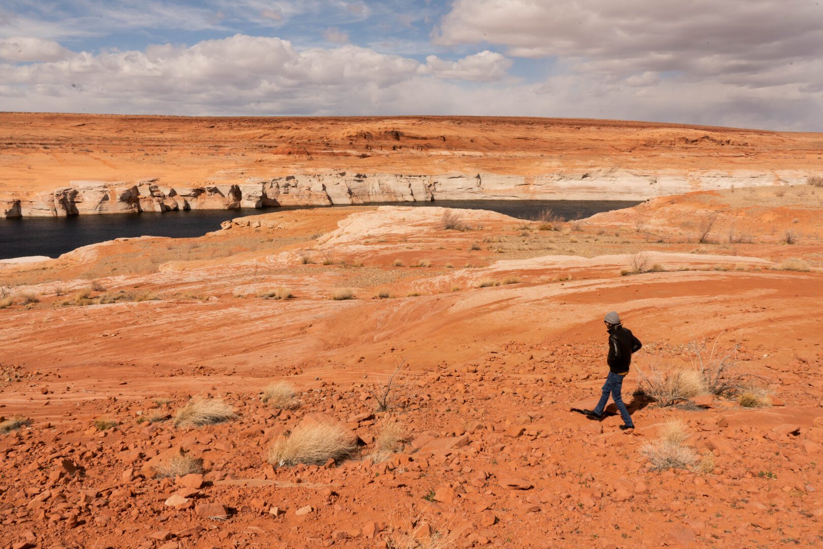 Glen Canyon Dam with an orange painted landscape and a crack in the earth that leads to the dam. A man in a jacket walks towards the scene, appearing like a figure.