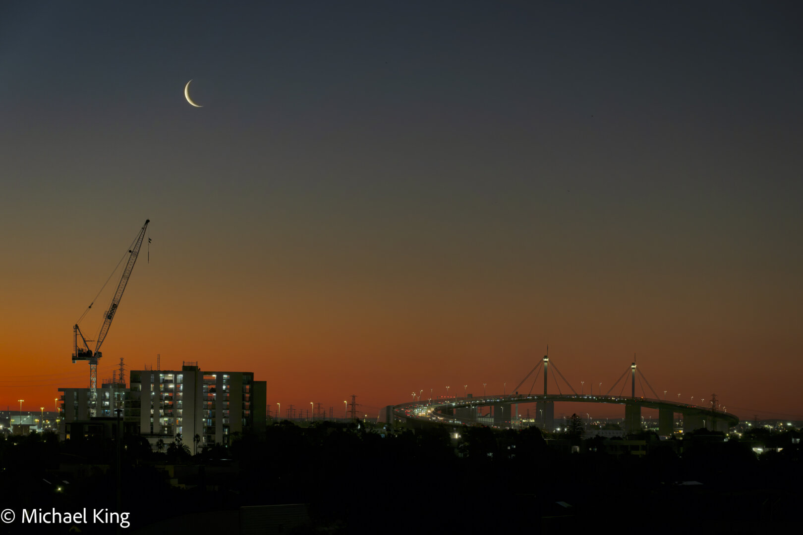 Bolte Bridge and a building under construction in silhouette against the sunset afterglow, offset by the setting crescent moon.