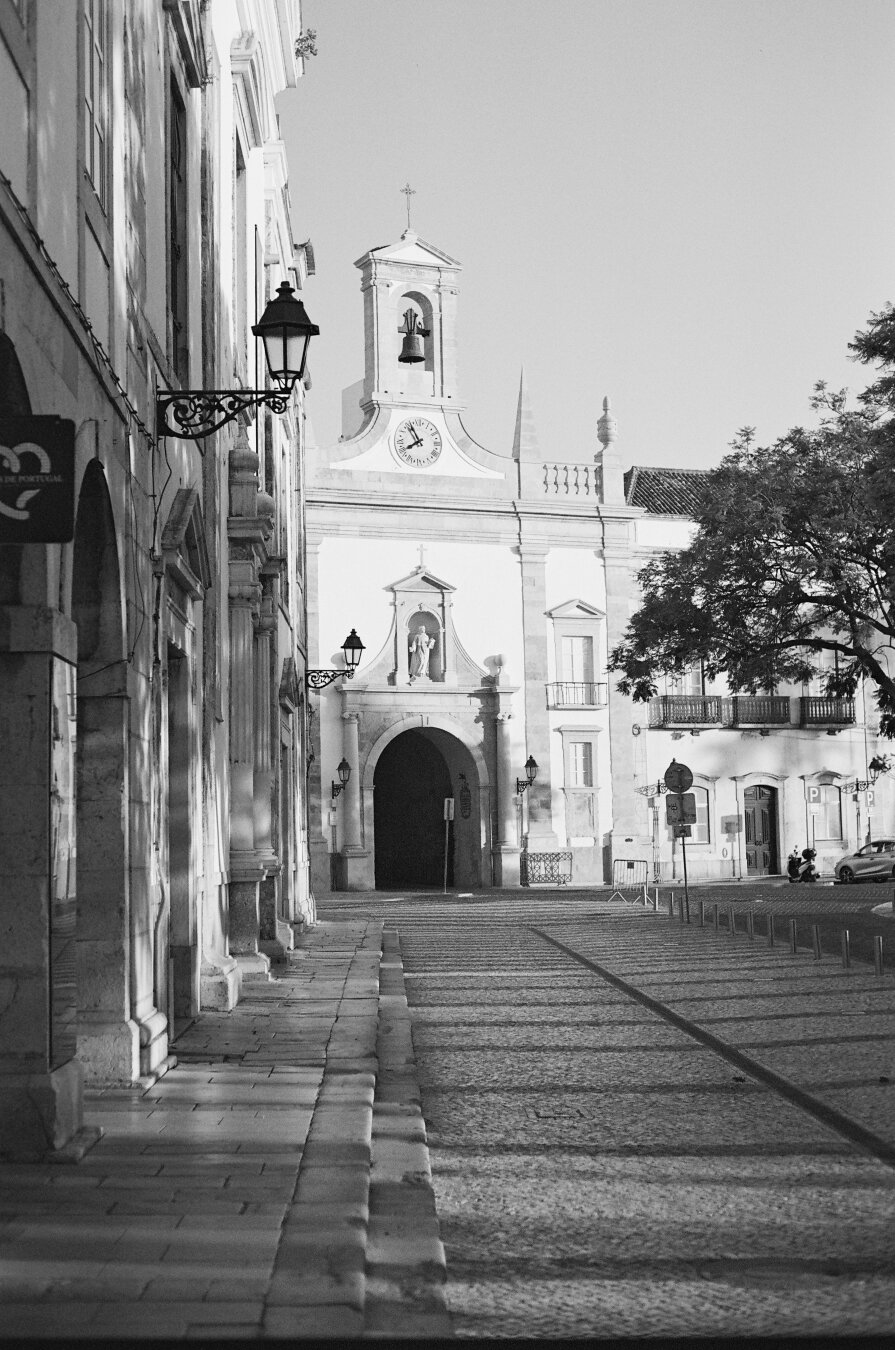 A black and white picture showing Faro's old town. At the centre of the frame is the gate of the old city from a square on the promenade.
Long shadows from right to left suggest the sun is low on the horizon.