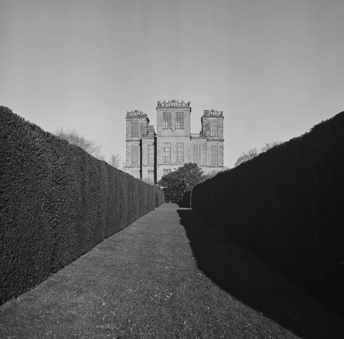 A black and white photograph of Hardwick Hall façade. The building stands at the end of a double hedged lawn path. The right hand side casts a dark shadow on the ground.