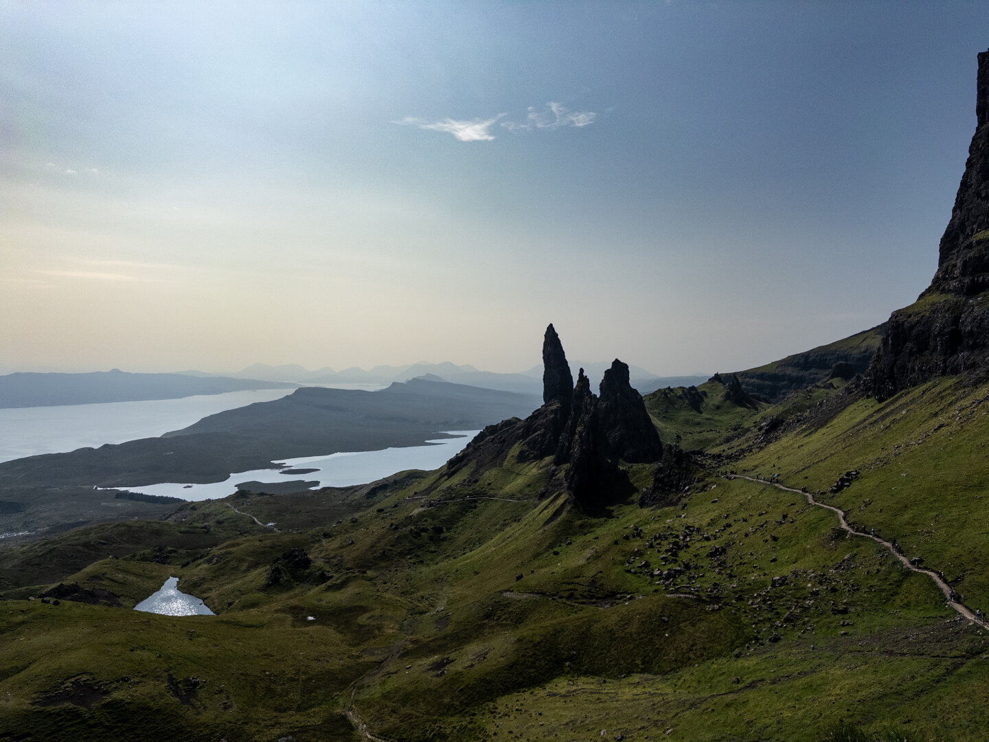A photo of The Old Man of Storr from the classic viewpoint. The fingers can be seen on the right side of the picture. The clear sky reveals a number of hazy hills in the background.
The picture is in landscape orientation