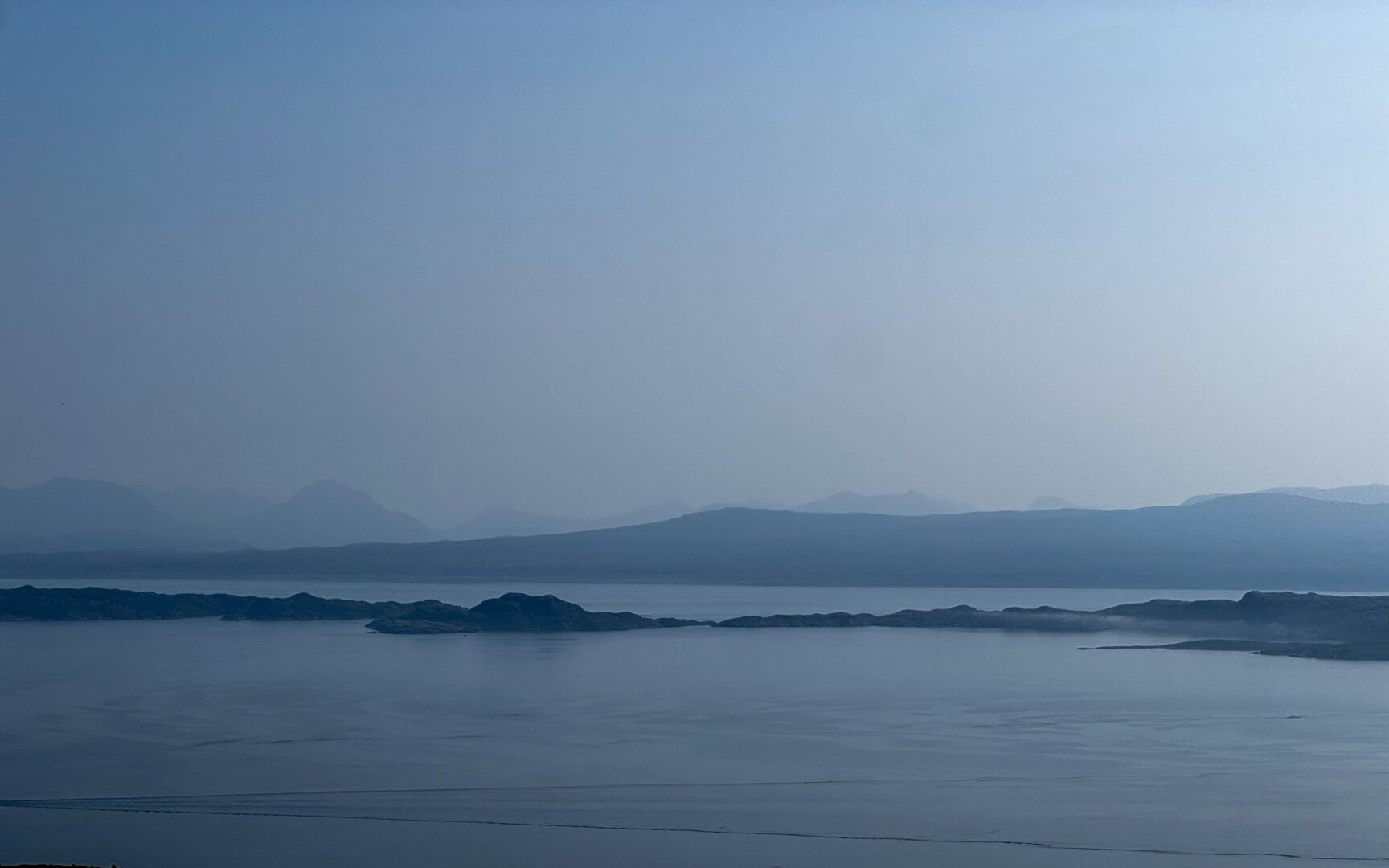 A view of Isle of Rona from The Storr. The island emerges from low morning clouds to a calm sea.
The wake of a single boat can be seen at the bottom of the picture.
The photo is in landscape orientation.