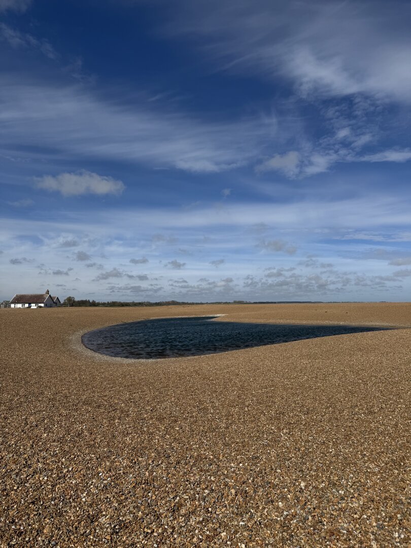 A picture of a small pool of water on a shingle beach. The pool has a droplet shape. In the background, on the left a white house. The photo is in portrait orientation.