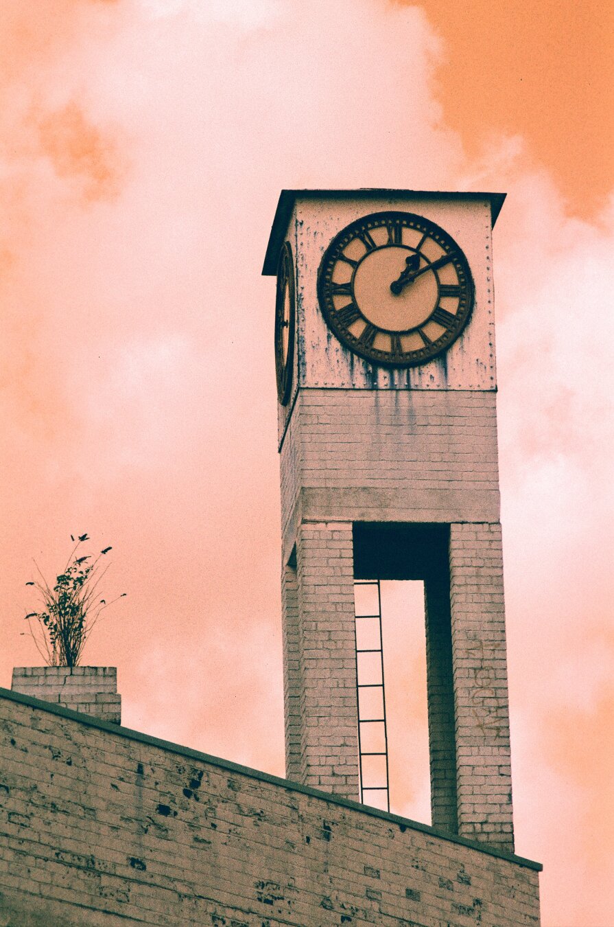 A photo of an old industrial clock tower in Bristol. The picture is shot on a film where colours are altered making the sky look red and the rest a pale azure. The clock tower has sharp corners and a light ladder can be seen at the back of the clock. The small tower tops a building where a bush-like plant has grown on top of