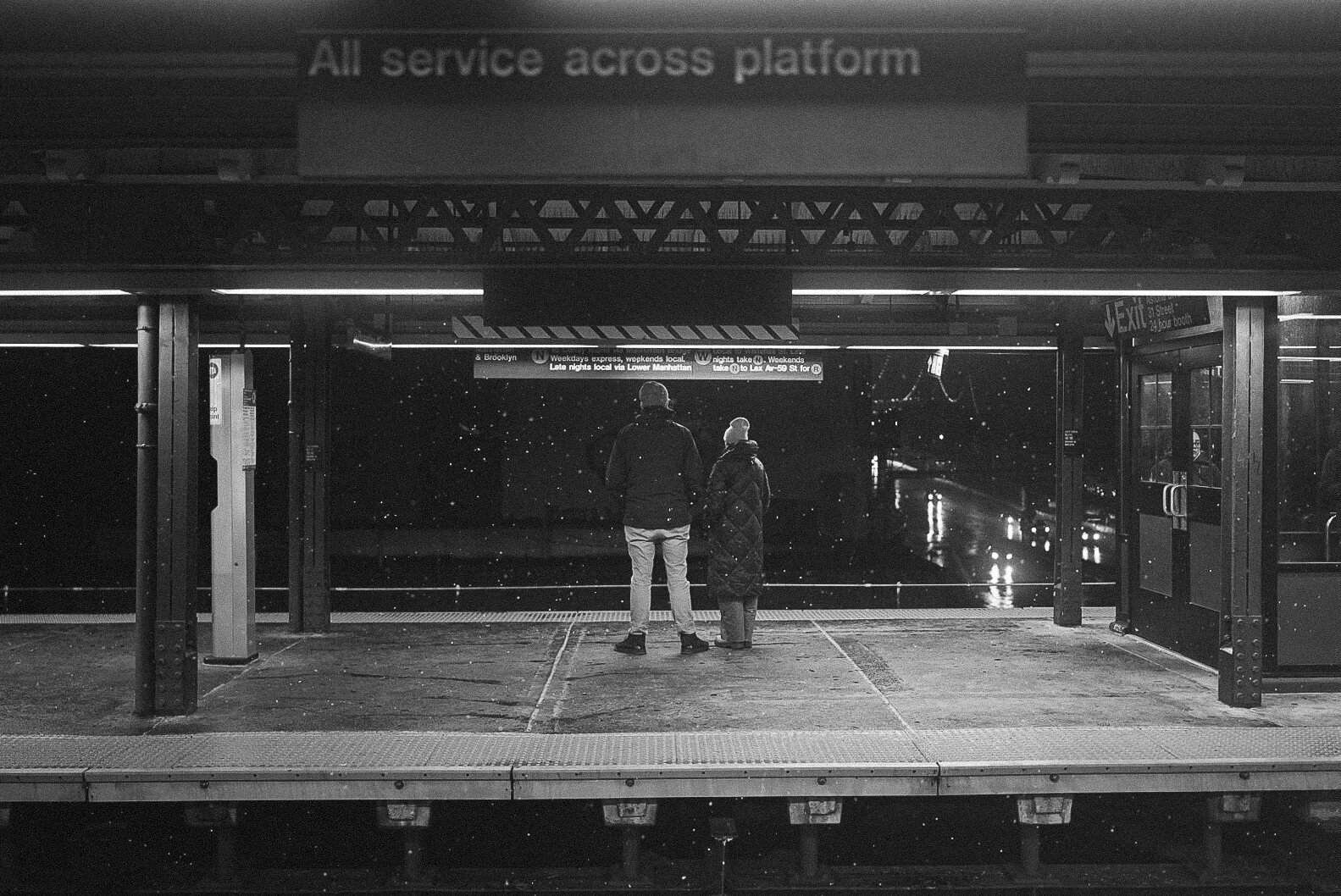 Two people waiting on a train platform during a light winter snow storm