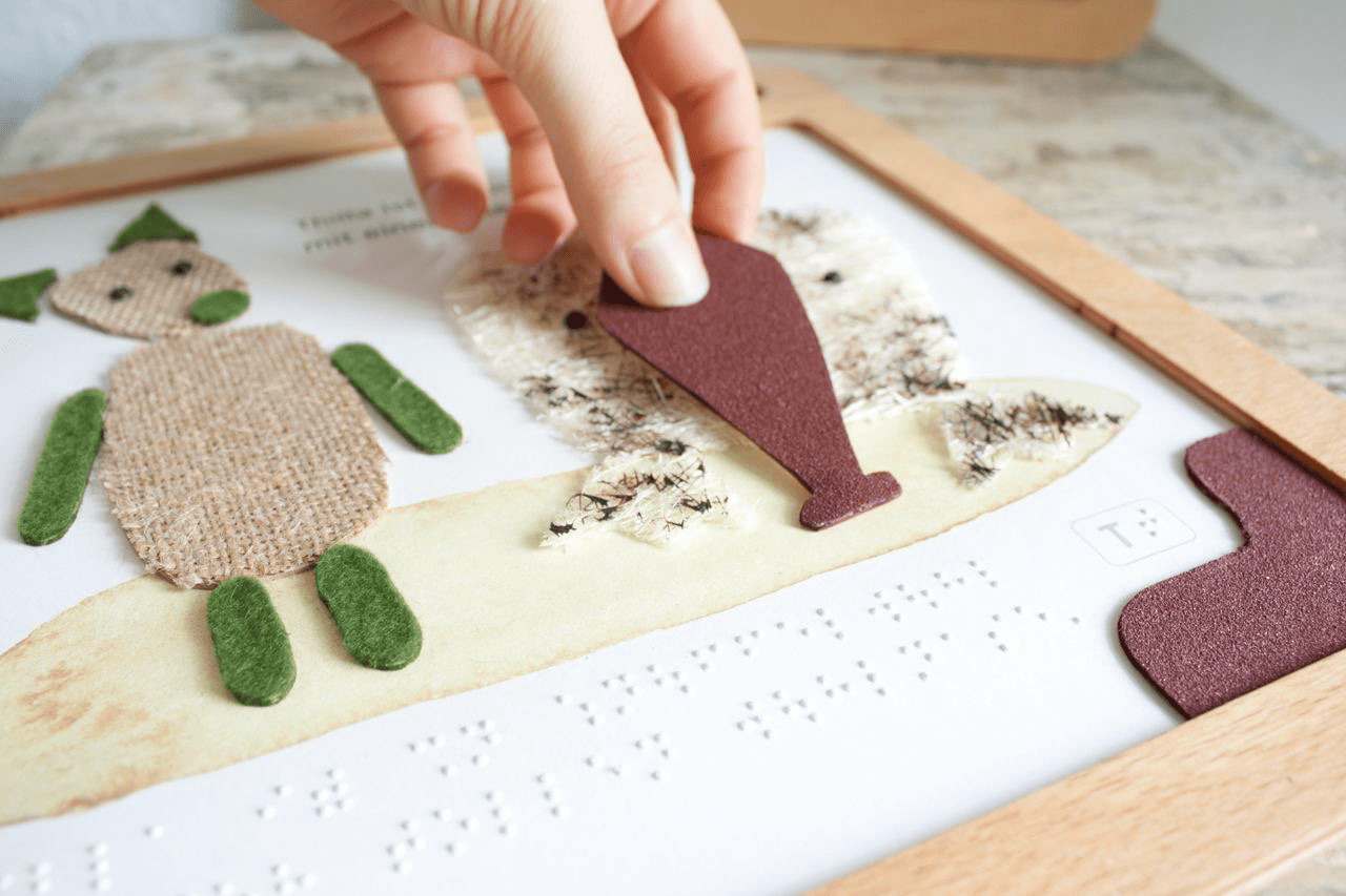 Eine Seite eines Holzbilderbuchs. Auf der Seite sind taktile Illustrationen, die bewegt werden können. Unten steht der Text in Braille, darüber in Schwarzschrift. Eine Hand puzzelt eins der Elemente auf eine Figur.