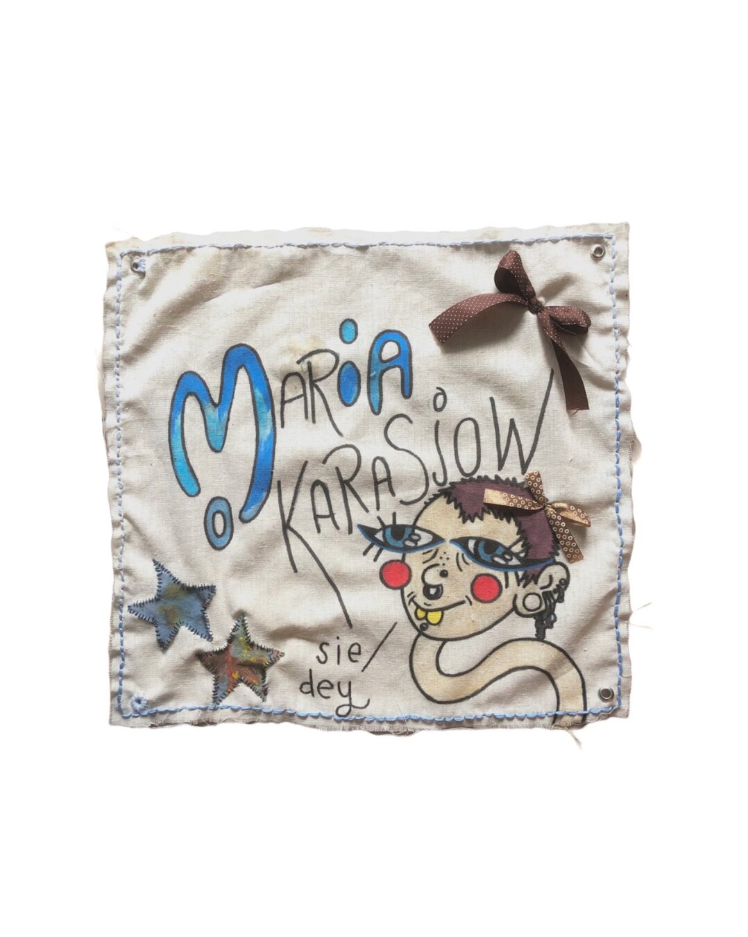 weißer Hintergrund mit einem kleinen quadratischen Banner aus hellem Stoff. Es wurden Sterne & Schleifen aufgenäht und eine Illustration, die die Künstlerin mit kurzen Haaren darstellen soll, eingezeichnet. In der Mitte steht der Name der Künstlerin: Maria Karasjow. Dabei sind das M-I-A hervorgehoben, um den Spitznamen Mia zu zeigen.