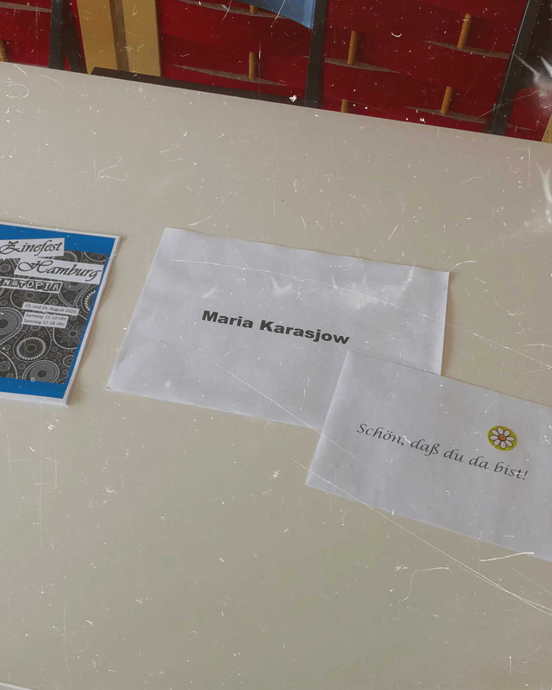 Auf einem Tisch liegen drei Zettel. Auf dem ersten steht: 2. Zinefest Hamburg ZINETOPIA. Auf dem zweiten steht: Maria Karasjow. Auf dem dritten: Schön, dass du da bist!