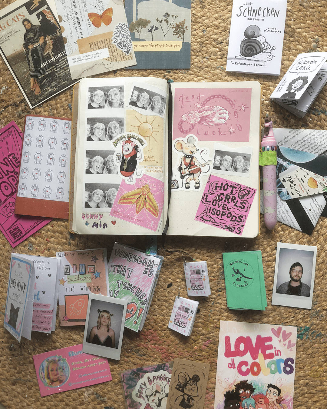 In der Mitte des Bildes liegt ein vollgeklebtes Notizbuch und drumzu liegen Zines, Postkarten und Sticker.