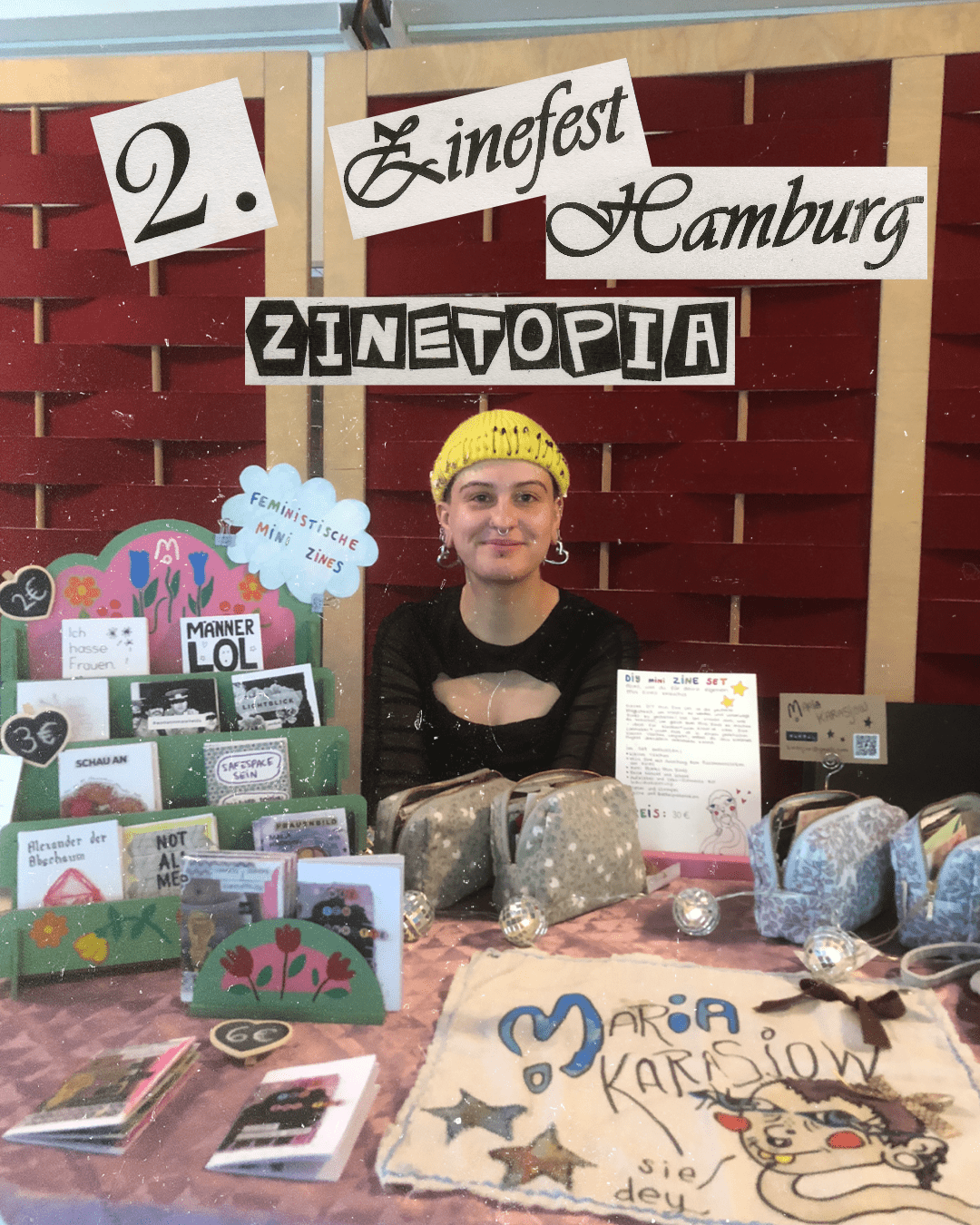 Eine Person (die Künstlerin des Accounts) sitzt hinter einem Tisch, der bedeckt mit Kunstartikeln ist. Über dey steht ein Text ais Papierschnipseln: 2. Zinefest Hamburg ZINETOPIA