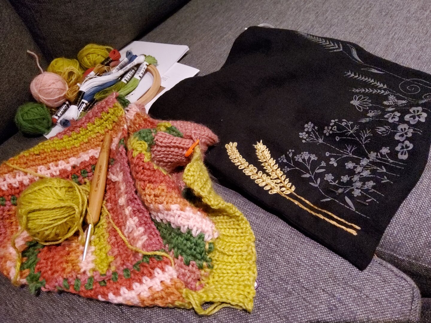Current WIPs: A multicolor crochet balaclava and an embroidery bag with yarn and tools on a grey sofa.