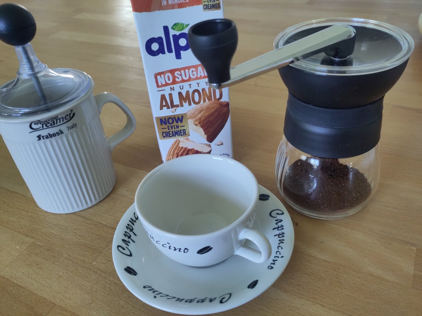 A light ceramic hand milk foamer, pack of almond milk, hand coffee grinder with coffee in the glass container and a white ceramic cup and saucer with the text "cappucino" flanked by coffee beans in black, on a wooden counter.