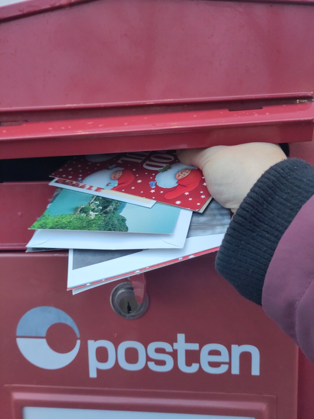 A hand putting several cards and letters in a red mailbox with the text Posten on it.
