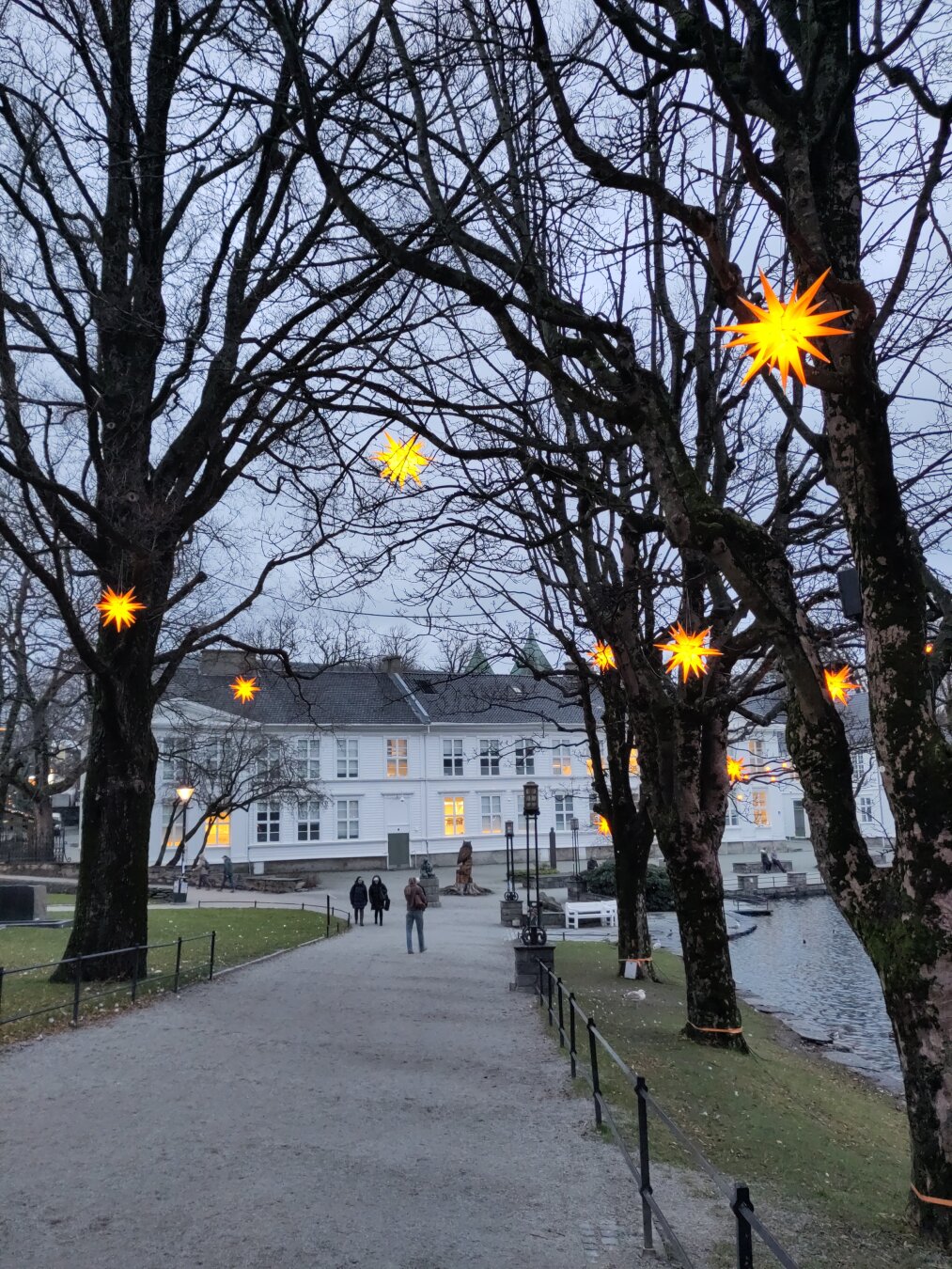 Står shaped lights hanging in trees that line a path along Byparken, the city park . A two-story white historical building with a black roof lies in the background.