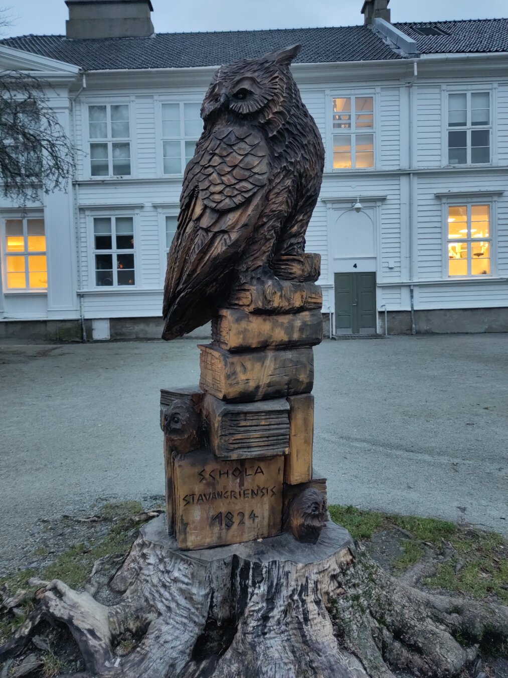 A wooden owl statue on a pile of books in front of a historical white building.