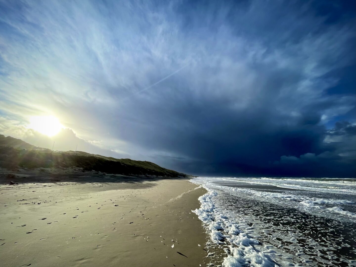 A view of Søndervig beach in Denmark with rolling waves, sun, blue skies and dark clouds.