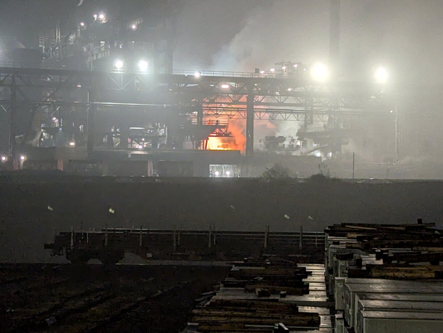 Im Zentrum des Bildes befindet sich ein großer, orangefarbener Lichtschein, der von einem Feuer oder glühendem Metall herrührt. Die Halle ist gefüllt mit verschiedenen Maschinen und Anlagen, die für die Stahlproduktion benötigt werden. Im Vordergrund des Bildes sind ein Güterwaggon mit Rohren und einige, nicht deutlich erkennbare, große Metallgegenstände zu sehen. Die Atmosphäre in der Halle wirkt heiß und gefährlich, was durch den intensiven Lichtschein und den dunklen Hintergrund noch verstärkt wird. Das Bild vermittelt einen Eindruck von der harten und gefährlichen Arbeit in einer Stahlgießerei.