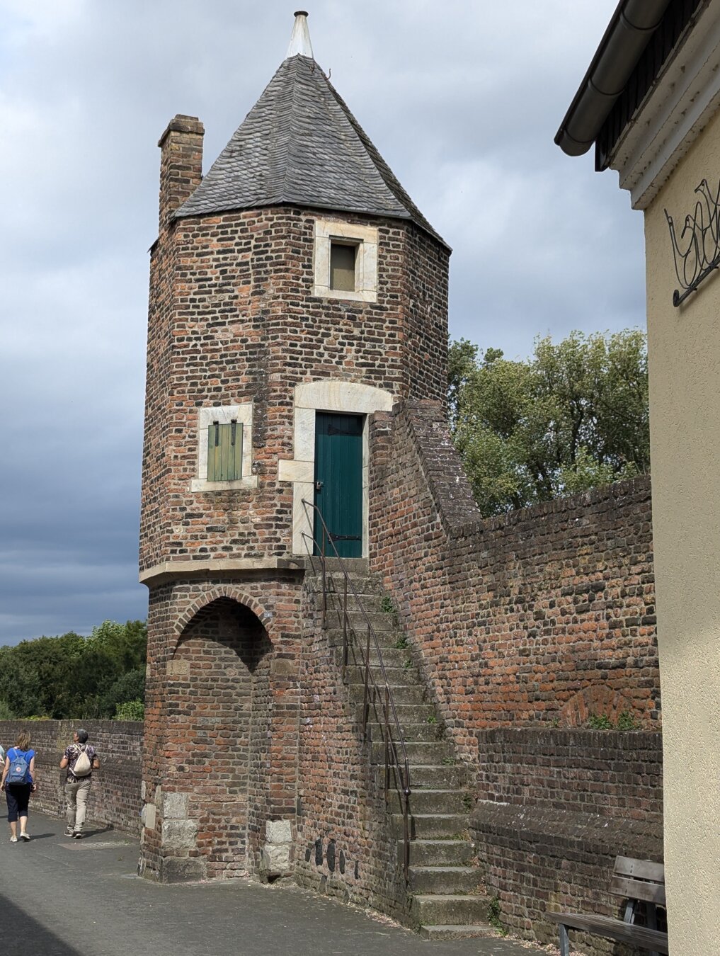 Das Foto zeigt einen alten, ziegelgemauerten Turm, eine sogenannte "Pfefferbüchse", mit einem spitzen Dach und einer kleinen Treppe, die zu einer Tür führt. Der Turm ist Teil der Stadtmauer der Feste Zons am Niederrhein.
Der Turm ist das Hauptelement des Bildes. Er ist aus roten Ziegelsteinen gebaut und hat ein spitzes, graues Dach. An der Vorderseite des Turms befindet sich eine kleine Treppe, die zu einer grünen Tür führt. Die Tür ist mit einem Vorhängeschloss versehen. Der Turm hat zwei kleine Fenster, eines auf jeder Seite der Tür. Die Fenster sind vergittert.

Der Turm ist von einer Mauer umgeben, die ebenfalls aus roten Ziegelsteinen besteht. Die Mauer ist niedrig und hat eine unregelmäßige Form. Hinter der Mauer befindet sich ein Baum mit grünen Blättern.