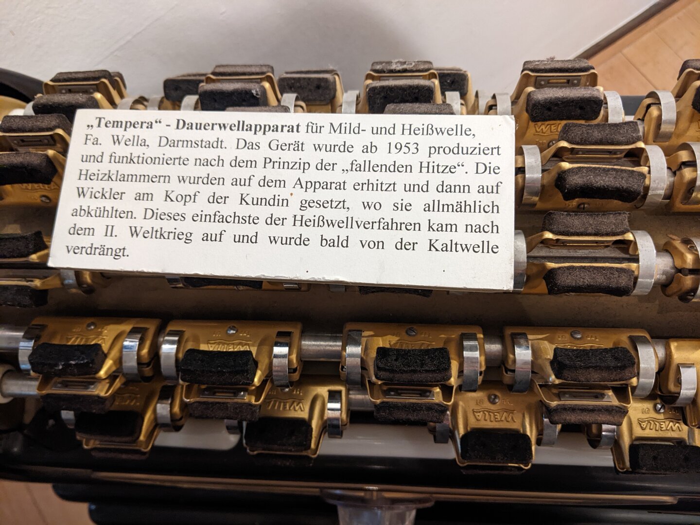 Das Bild zeigt einen „Tempera" - Dauerwellapparat für Mild- und Heißwelle, hergestellt von der Firma Wella in Darmstadt. 
Der Apparat funktionierte nach dem Prinzip der „fallenden Hitze". Das bedeutet, dass die Heizklammern auf dem Apparat erhitzt und dann auf Wickler am Kopf der Kundin gesetzt wurden, wo sie allmählich abkühlten. Dieses Verfahren war das einfachste der Heißwellverfahren und kam nach dem Zweiten Weltkrieg auf den Markt. 
Auf dem Bild ist eine Reihe von Heizklammern zu sehen, die auf dem Apparat aufgereiht sind. Die Klammern sind aus Metall gefertigt und haben eine spezielle Form, die es ermöglicht, sie auf den Wicklern am Kopf der Kundin zu befestigen. Der Apparat selbst ist aus einem hellen Material gefertigt und trägt den Schriftzug „WELLA" in großen Buchstaben. Der Apparat selbst ist aus einem hellen Material gefertigt und trägt den Schriftzug „WELLA" in großen Buchstaben. Darunter befindet sich ein kleineres Schild mit der Aufschrift „Tempera" - Dauerwellapparat"