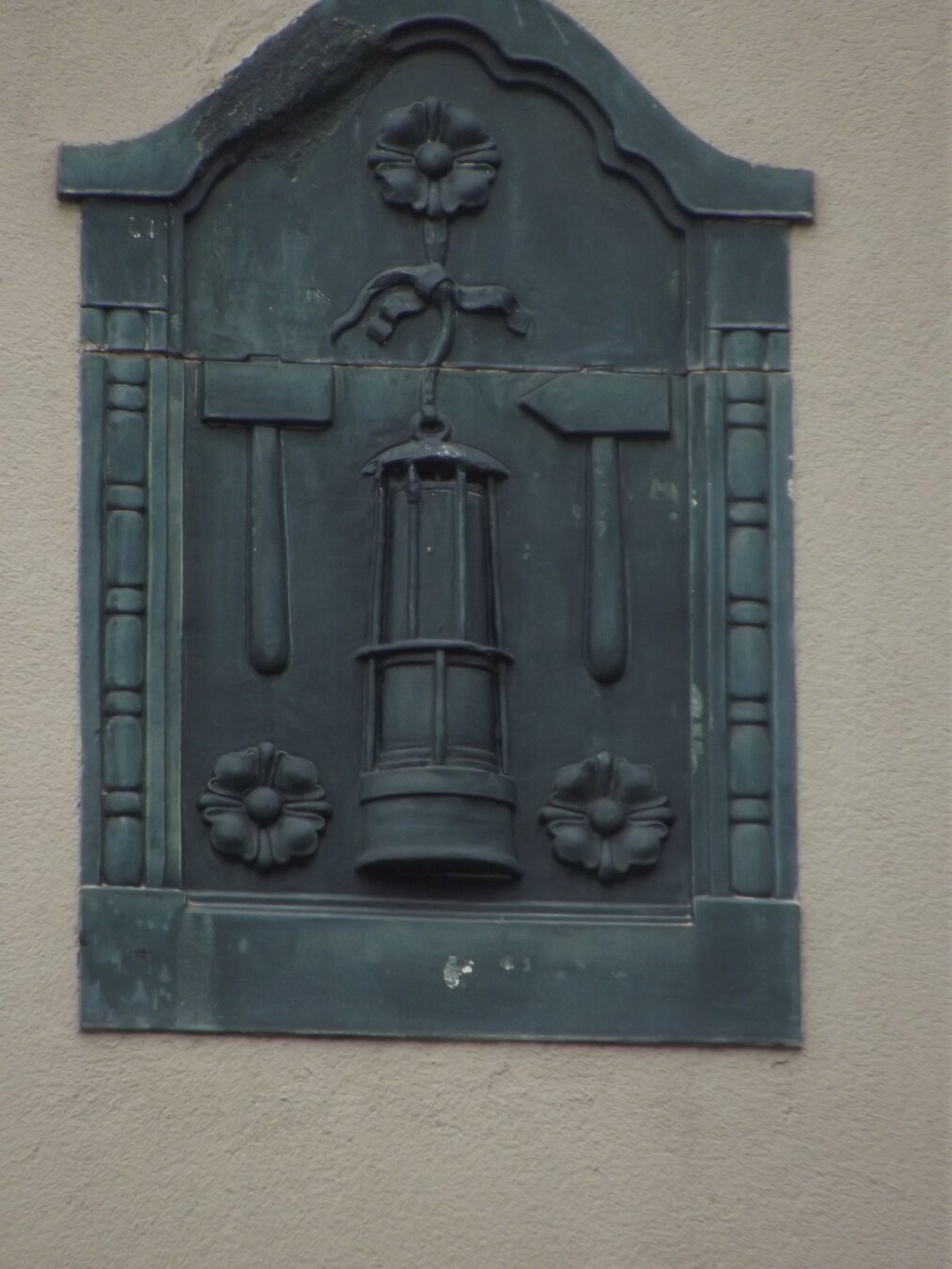 Das Bild zeigt ein Relief in einer rechteckigen, oben gewölbten Form.
Es besteht  aus einem dunklen Material, möglicherweise Metall und ist an einer helleren Wand angebracht.
Im Zentrum des Reliefs hängt eine Grubenlampe. 
 Links und rechts neben der Lampe sind Hammer und Schlegel angebracht. Sie sind symmetrisch angeordnet und zeigen nach oben.
Oben und unten an den Hämmern sind jeweils stilisierte Blumen angebracht. Sie haben eine einfache Form mit mehreren Blütenblättern.
Das Relief ist von einem einfachen Rahmen umgeben. Links und rechts sind Säulen angebracht, während oben und unten jeweils eine horizontale Linie den Rahmen abschließt.