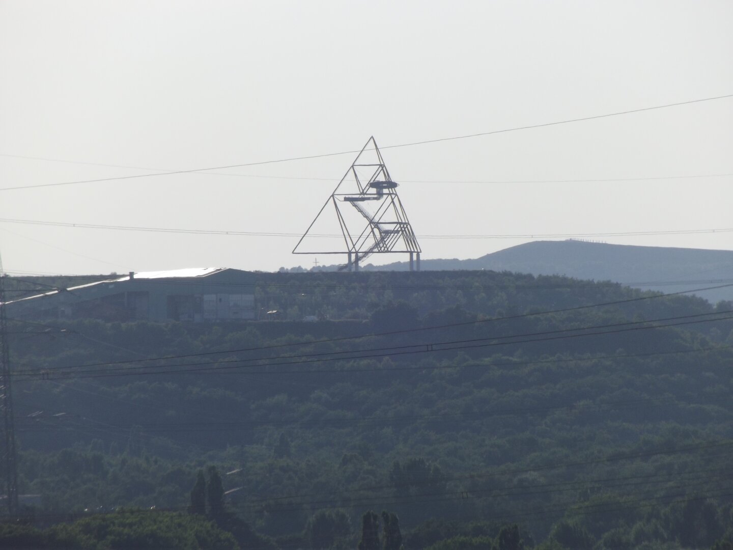 Dieses Bild zeigt eine Außenaufnahme des Tetraeders im Quadrat Bottrop, einer markanten Landmarke im Ruhrgebiet. Der Tetraeder ist ein großes, pyramidenförmiges Bauwerk mit einer Stahlkonstruktion, die ein dreidimensionales Gittermuster bildet.
 Das Hauptaugenmerk des Bildes ist der Tetraeder. Er ist in der Mitte des Bildes platziert und ragt hoch in den Himmel. Seine Stahlkonstruktion ist gut sichtbar und verleiht ihm ein leichtes, transparentes Aussehen. Die Struktur besteht aus mehreren diagonalen Linien, die ein dreieckiges Muster bilden.
Der Tetraeder befindet sich auf einer Anhöhe. Unterhalb des Tetraeders sind Bäume und Sträucher zu sehen, die einen grünen Hügel bilden. Im Hintergrund befindet sich eine weitere Anhöhe, die ebenfalls von Bäumen bedeckt ist. Der Himmel ist hell und leicht bewölkt, was einen neutralen Hintergrund für den Tetraeder bildet.