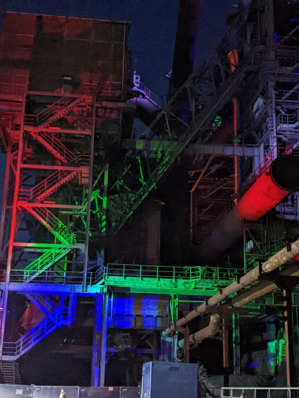 Das Foto zeigt eine nächtliche Außenaufnahme eines Industriegeländes, das durch bunte Lichter beleuchtet wird. Im Vordergrund befindet sich ein großer, dunkler Kasten, der möglicherweise ein Gebäude oder eine Maschine darstellt.
Im Hintergrund sind mehrere hohe, schlanke Strukturen zu sehen, die wie Schornsteine oder Fördertürme aussehen. Diese sind ebenfalls dunkel, aber ihre Umrisse sind durch die Beleuchtung erkennbar.
Ein markantes Merkmal des Bildes ist die farbige Beleuchtung. Das gesamte Gelände ist in ein diffuses, buntes Licht getaucht, das von verschiedenen Scheinwerfern oder Lichtquellen zu stammen scheint. Die Farben wechseln zwischen Rot, Grün, Blau und Gelb, wodurch eine lebendige und dynamische Atmosphäre entsteht.
Der Himmel über dem Industriegelände ist dunkel und wolkenverhangen. Es sind keine Sterne oder anderen Himmelskörper zu erkennen.