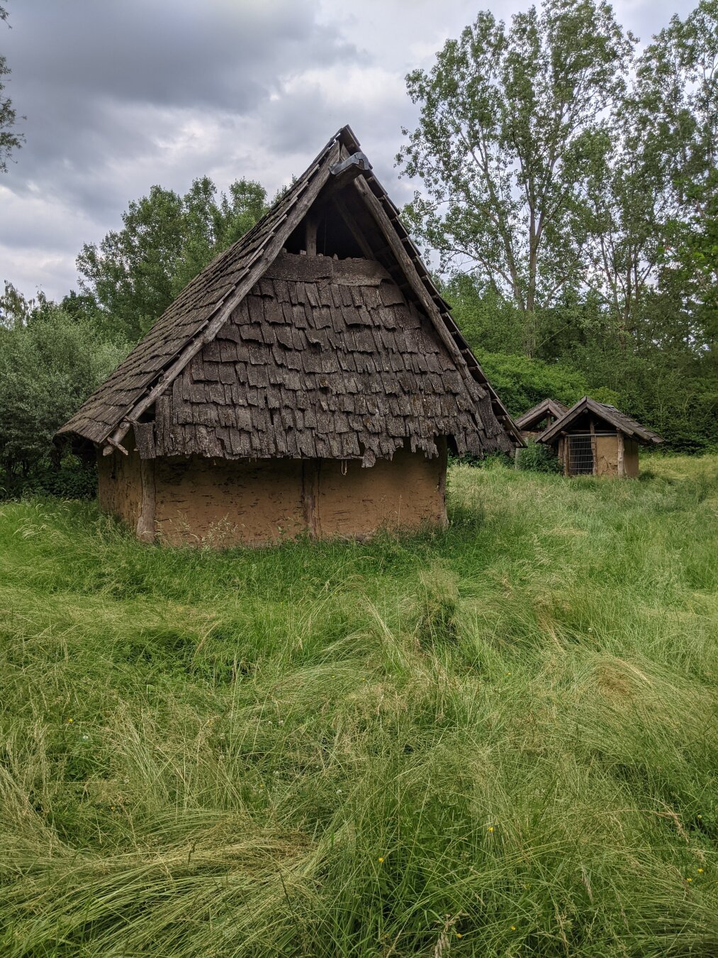 Das Bild zeigt drei Hütten mit Wänden aus gelbem Lehm, die Dächer sind spitz und aus Baumstämmen und Holzschindeln. Bei den beiden hinteren, die dicht beieinander stehen, sieht man, dass das Dach nur an den Seiten geschlossen ist. Das vordere hat eine Türöffnung, die allerdings mit einem Gitter verschlossen ist. Das Dach des vorderen Gebäudes ist im oberen Teil offen. Die Hütten weisen teils stärkere Beschädigungen auf.
Rings um die Hütten ist hohes Gras, im Hintergrund wachsen Bäume. Der Himmel ist bewölkt.