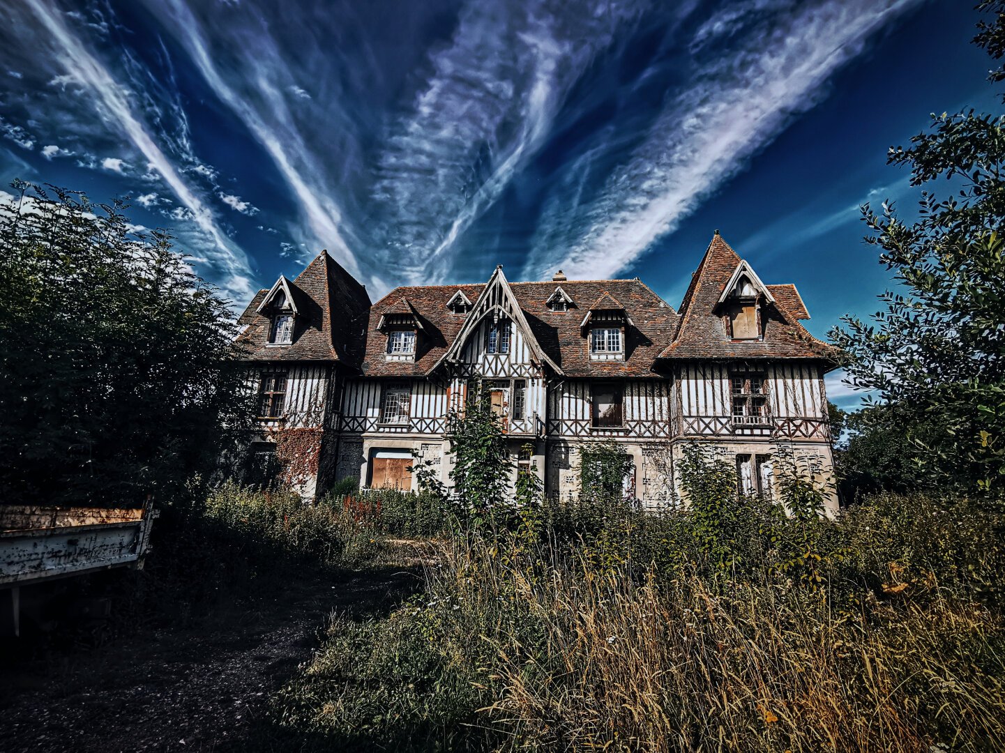 A colombage mansion in an overgrown garden, forgotten and abandoned for long, under a cloudy sky.