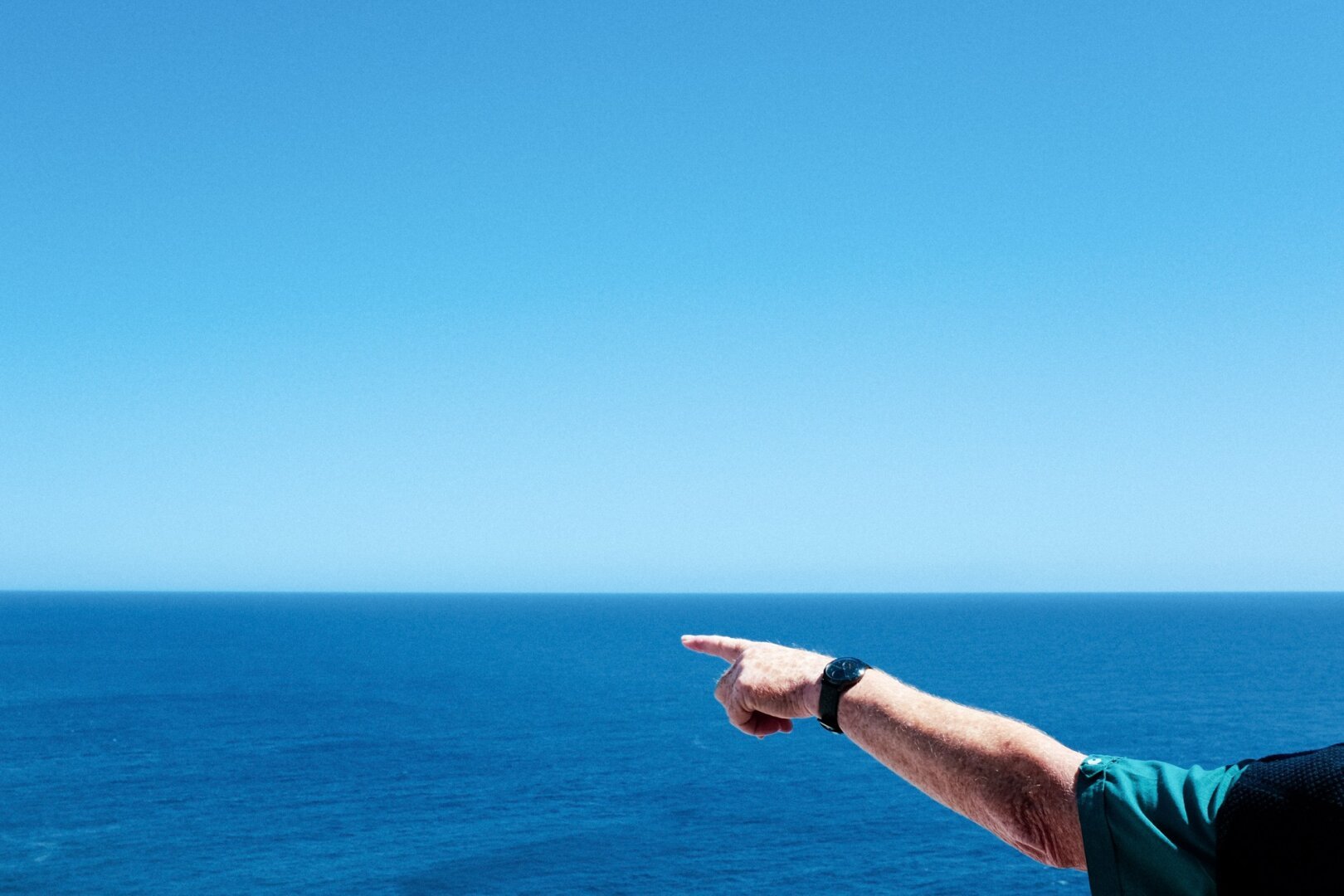 Das Meer und der blaue Himmel. Davor ein Arm mit Armbanduhr, der Zeigefinger zeigt in die Luft.