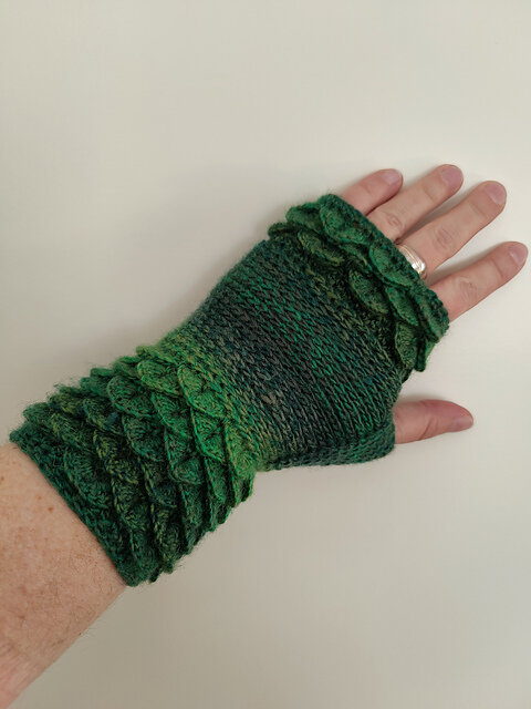 A left hand wearing a fingerless glove crocheted in a gradient of green tones, ranging from deep forest green to bright emerald. The glove features a prominent scale-like texture across the wrist and knuckles, resembling dragon or reptile skin. The fingers are exposed, and the wearer has a silver ring on the ring finger. The craftsmanship highlights intricate stitchwork and a fantasy-inspired design.