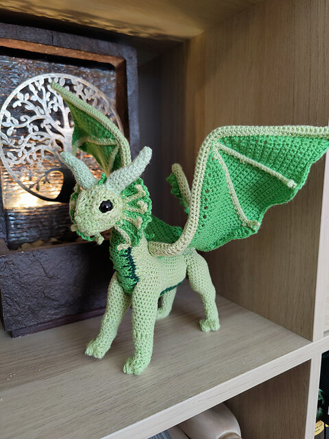 Crocheted dragon figurine posed on a wooden shelf, made with light and dark green yarn. The dragon features large, structured wings with a dark green ridge, a textured body, and expressive black eyes. Its face is detailed with protruding ears and a frilled neck, evoking a fantasy creature. Behind it, a decorative panel with a tree design adds a mystical backdrop, enhancing the handcrafted, whimsical atmosphere.