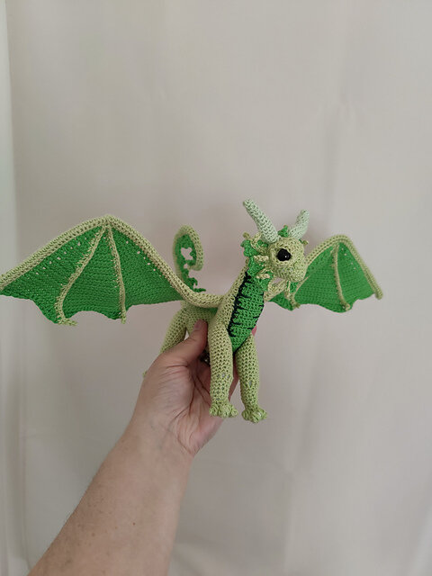 Hand holding a crocheted dragon made entirely from various shades of green yarn. The dragon features detailed wings, a curled tail, and prominent eyes. The craftsmanship showcases intricate shaping and attention to detail, creating a whimsical, handmade interpretation of a mythical creature.